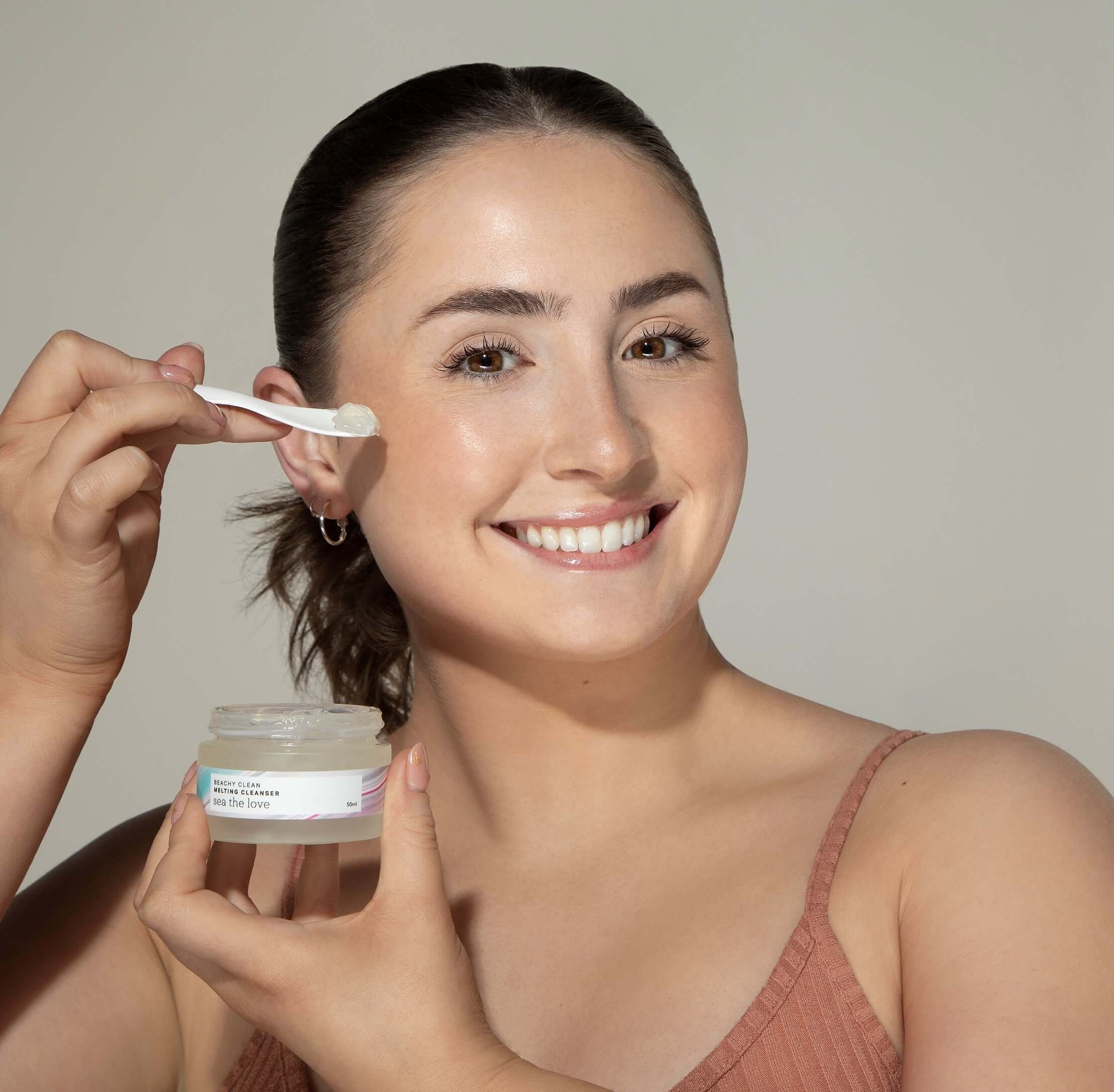 Woman holding a jar of Beachy Clean Melting Cleanser in one hand, while holding a spatula with a scoop of the gel cleanser in her other hand.