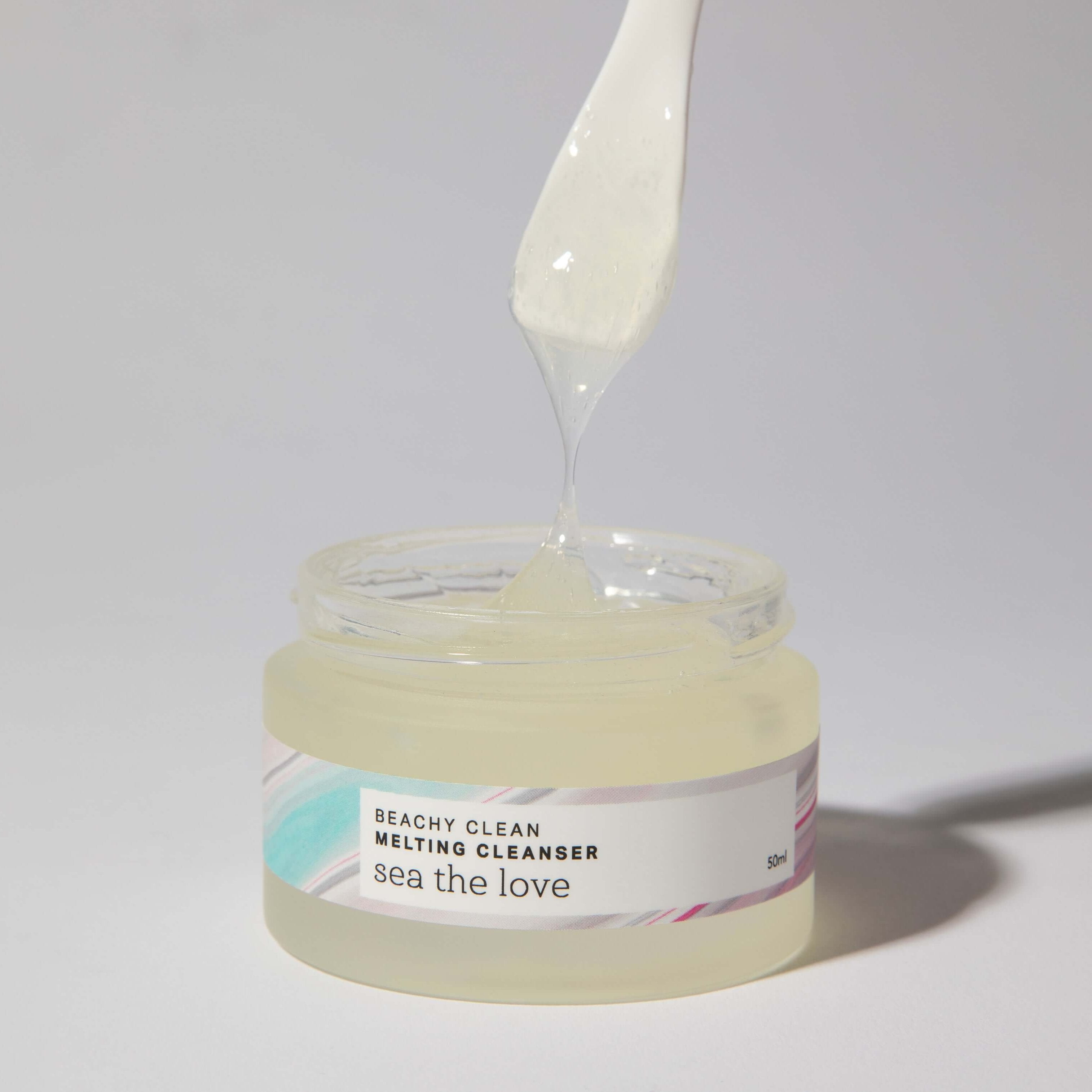An open jar of the Beachy Clean Melting Cleanser from Sea the Love, with a spatula taking a scoop of the product showcasing the cleansers gel consistency