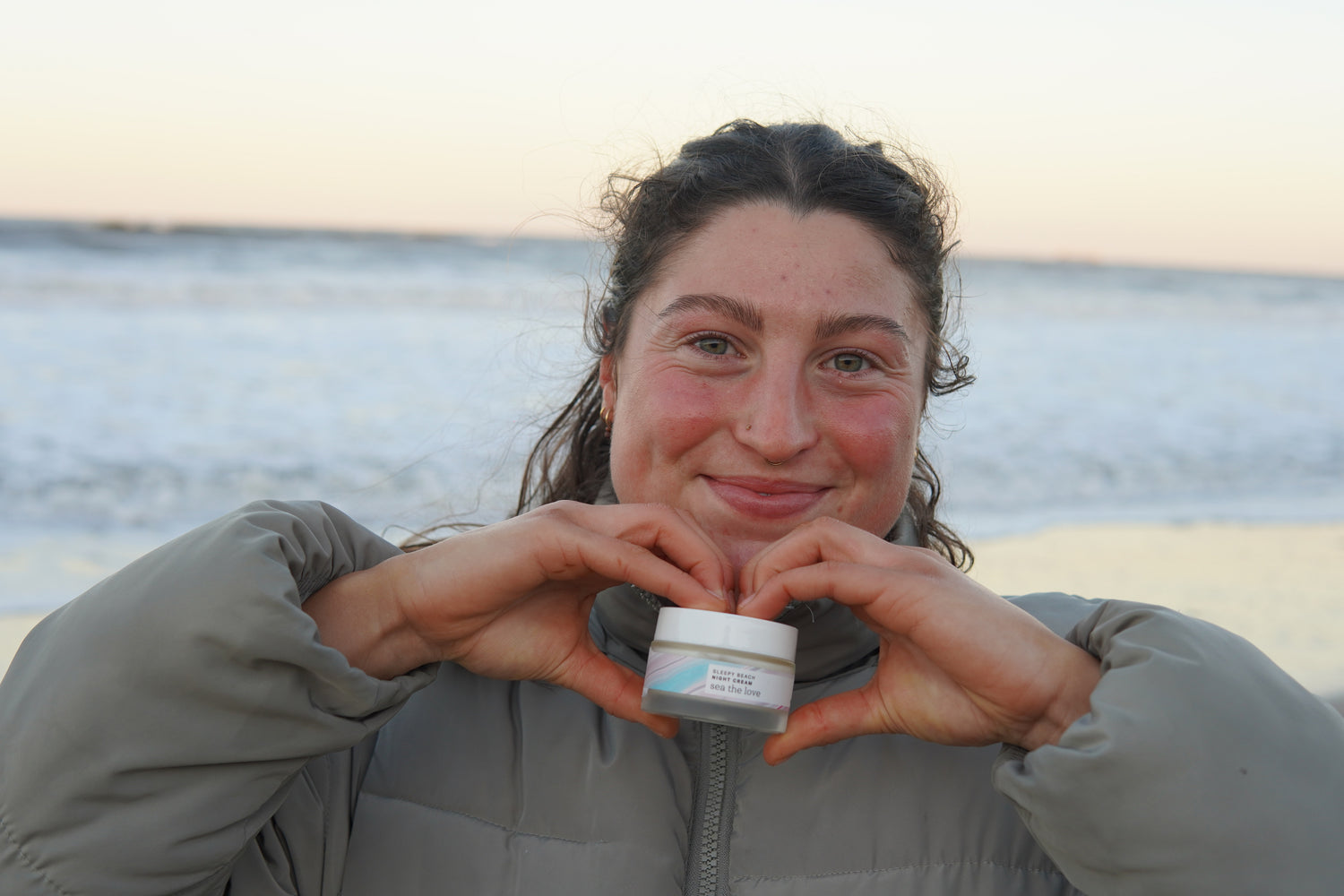 woman smiling holding a sea the love night cream, with the sea behind her