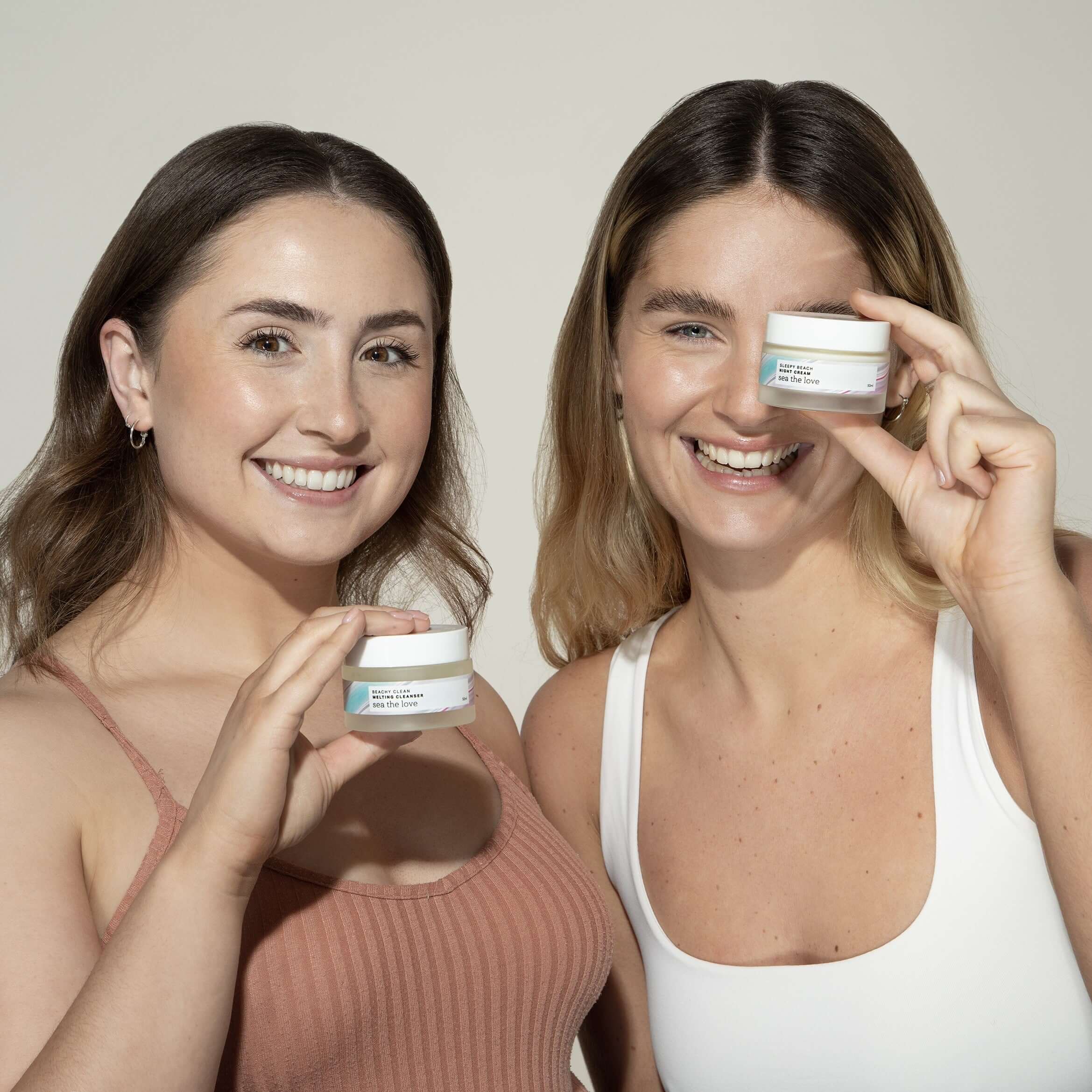 Two women holding up a Sea the Love ocean inspired skincare product, one with beachy clean melting cleanser, the other with sleepy beach night cream.