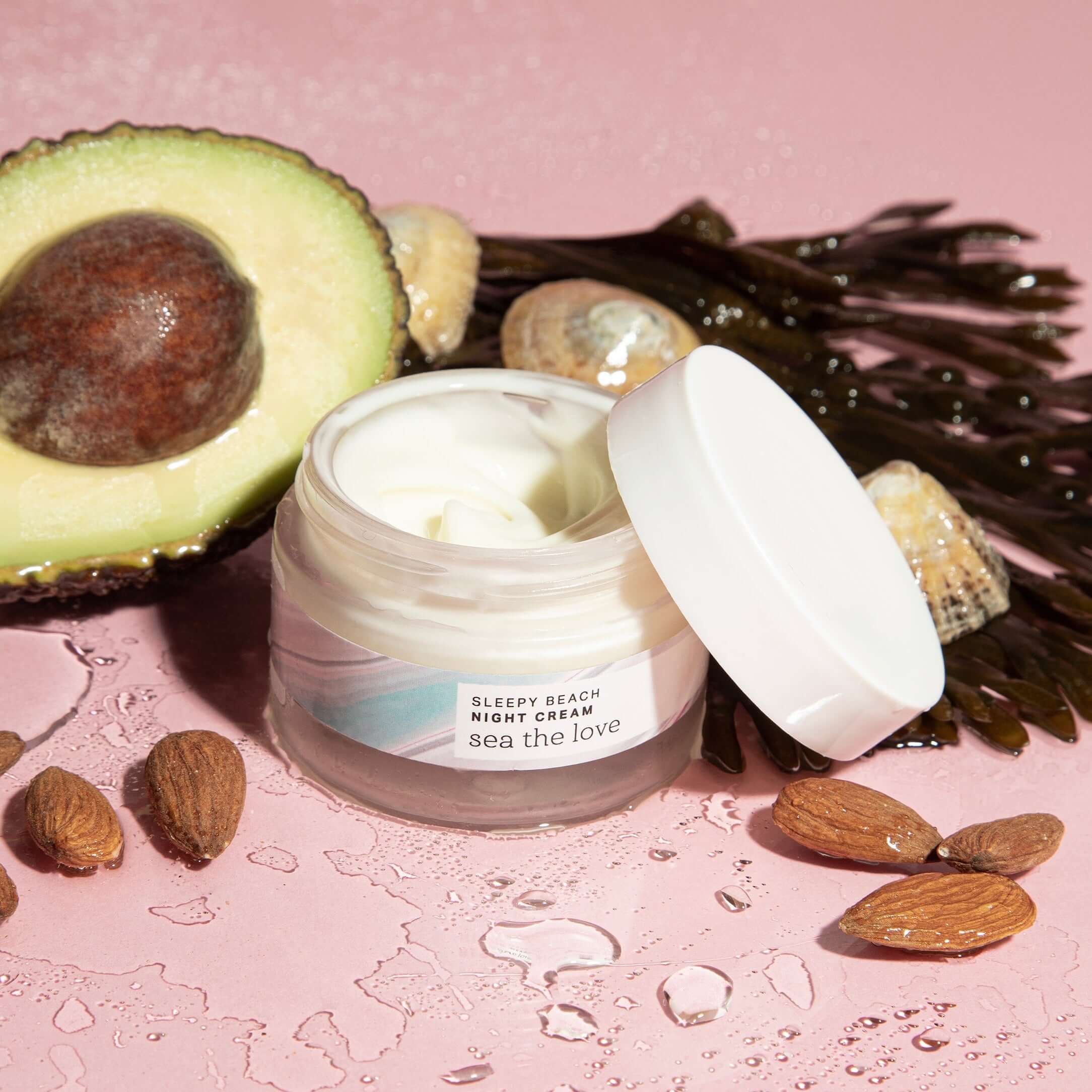 A jar of Sleepy Beach Night Cream surrounded by natural skincare ingredients including an avocado, almonds, water and seaweed.