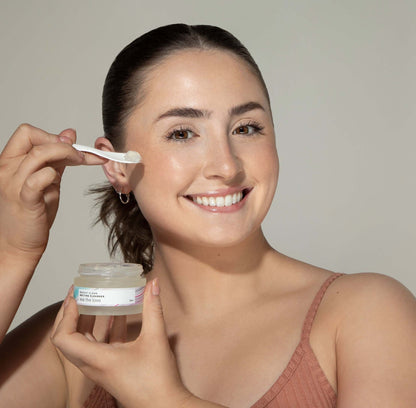 Woman holding a jar of Beachy Clean Melting Cleanser in one hand, while holding a spatula with a scoop of the gel cleanser in her other hand. 