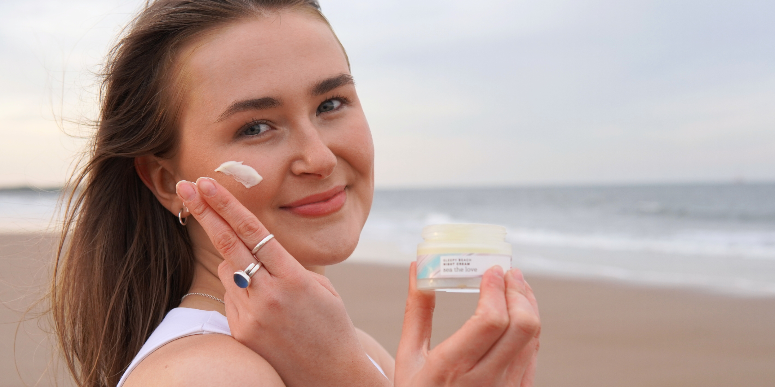 A woman on the beach holding a jar of night cream in one hand, while switching the moisturiser on her cheek,