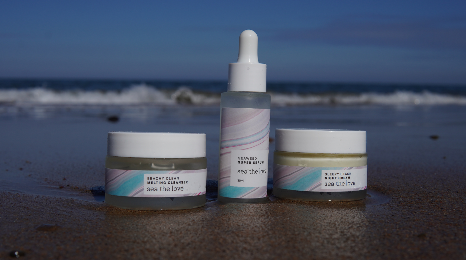 a cleanser, serum, and night cream, lined up on the shoreline with the sea crashing in the background