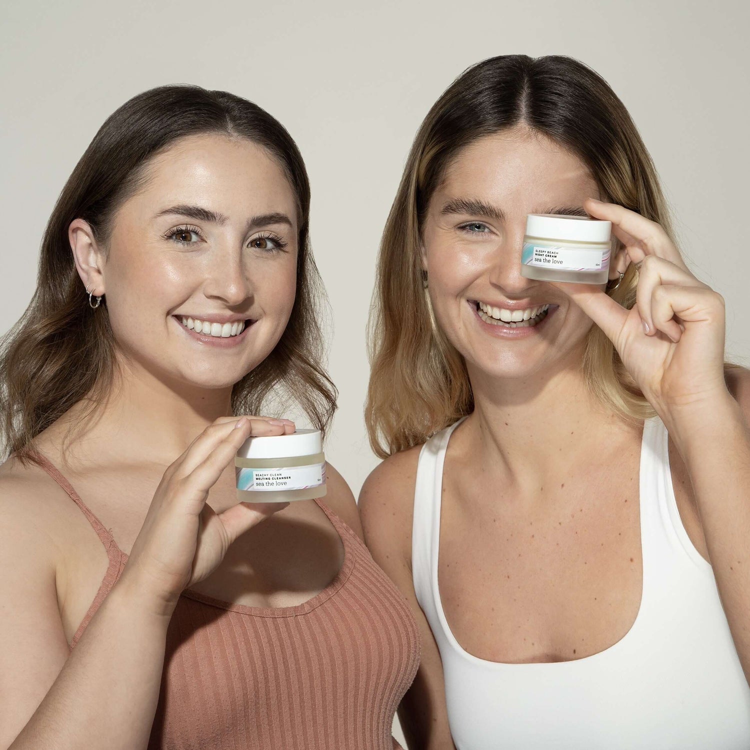  Two women holding up a Sea the Love ocean inspired skincare product, one with beachy clean melting cleanser, the other with sleepy beach night cream.