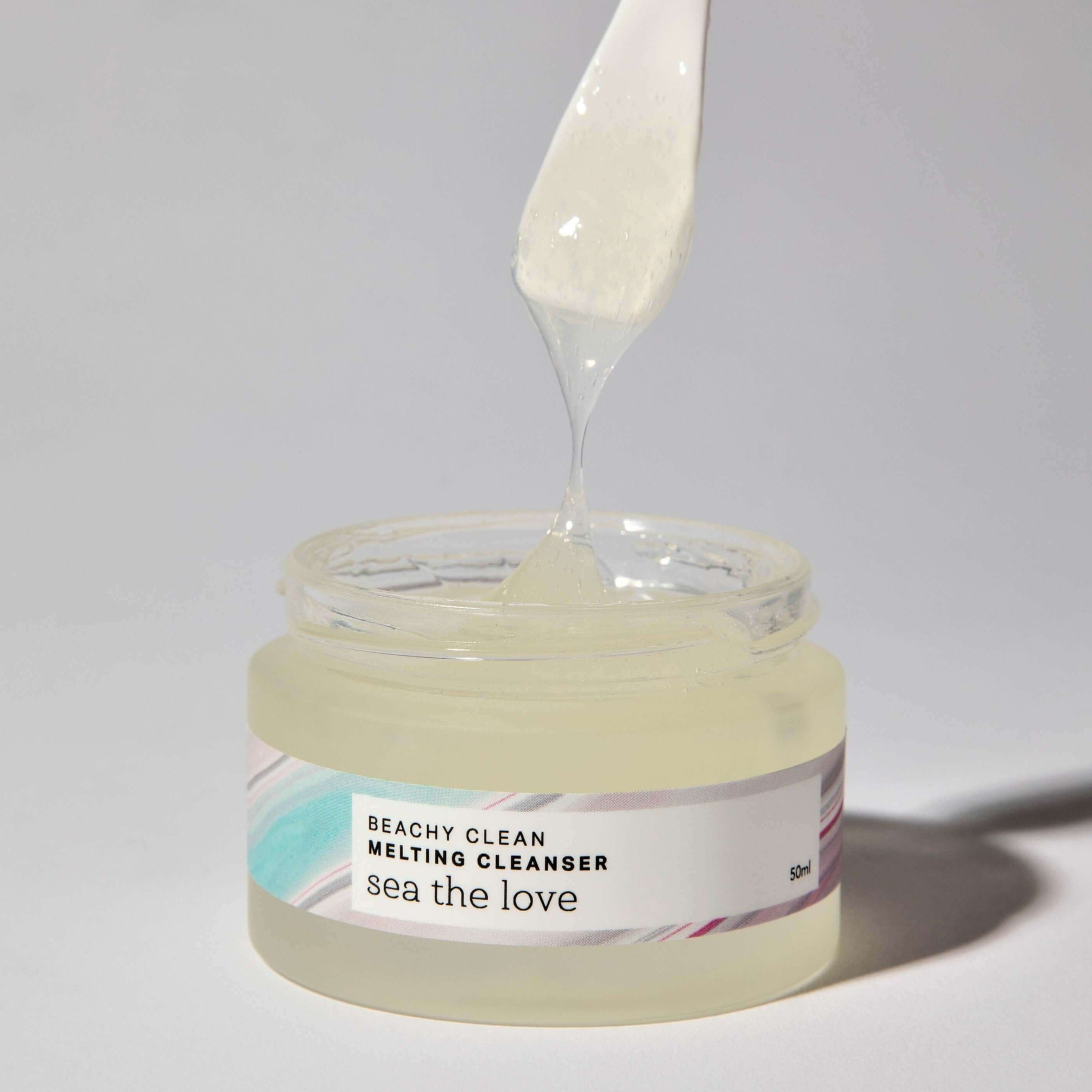 An open jar of the Beachy Clean Melting Cleanser from Sea the Love, with a spatula taking a scoop of the product showcasing the cleansers gel consistency 