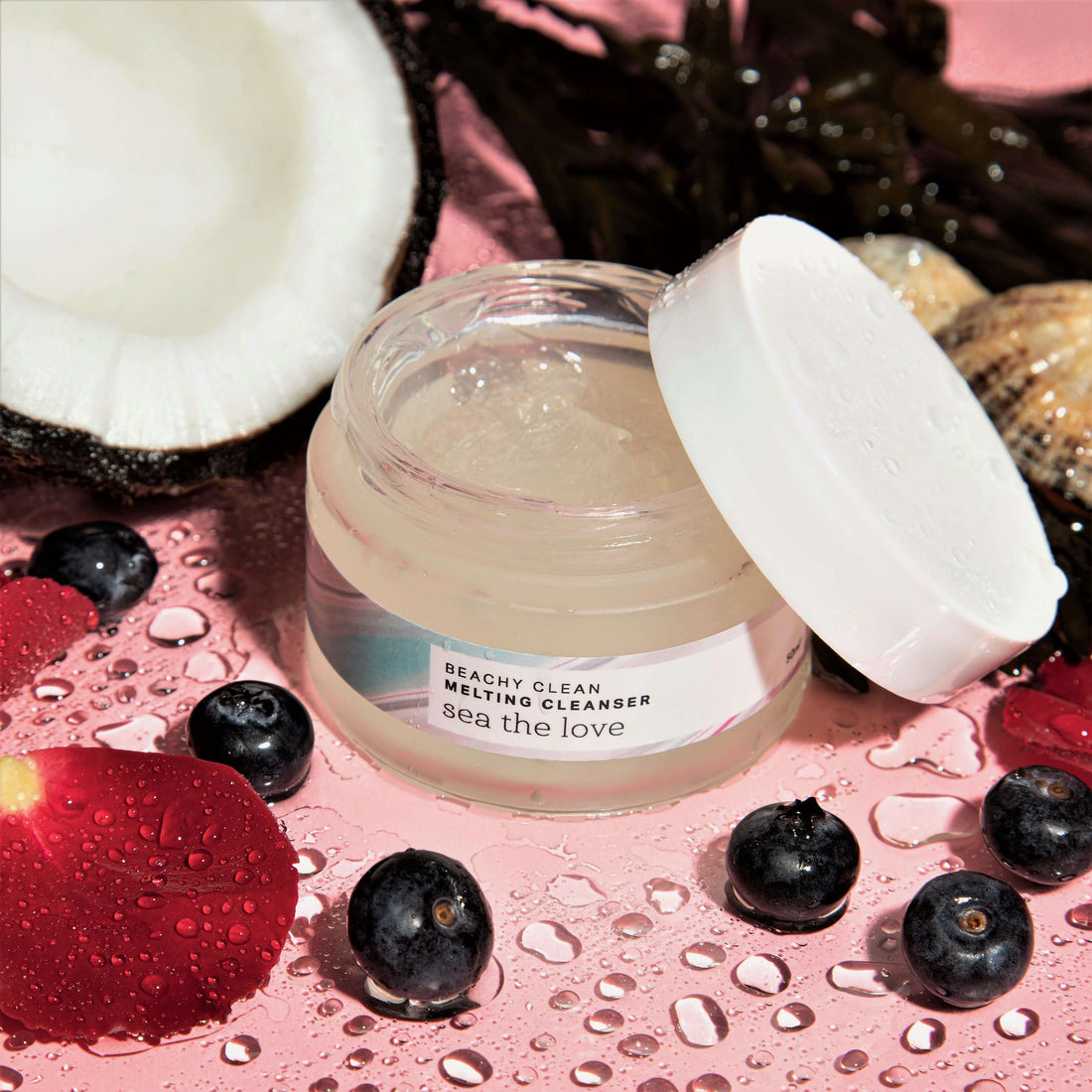 A jar of Beachy Clean Melting Cleanser surrounded by natural skincare ingredients including blueberries, coconuts, and seaweed. 